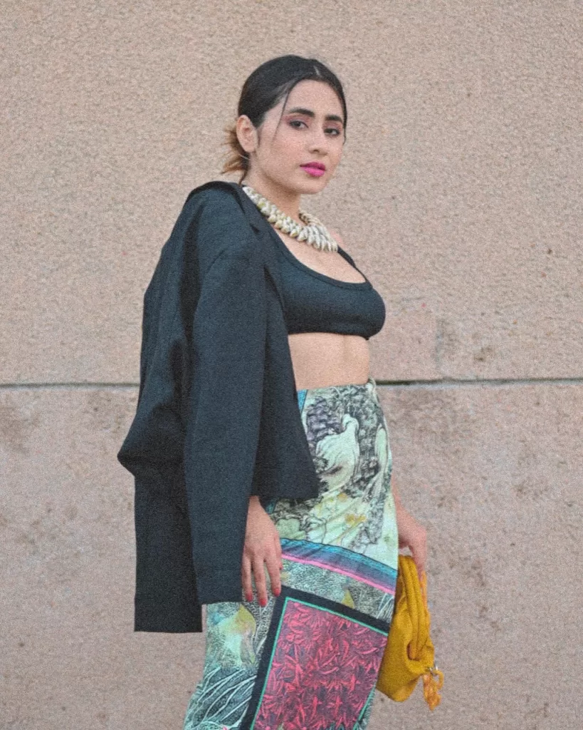 in a tailored black blazer and vibrant digital print tie-up skirt, confidently looking over her shoulder, holding a mustard-yellow bag. Paired with sleek ankle boots, her ensemble exudes a mix of power and playful chic against a neutral backdrop.