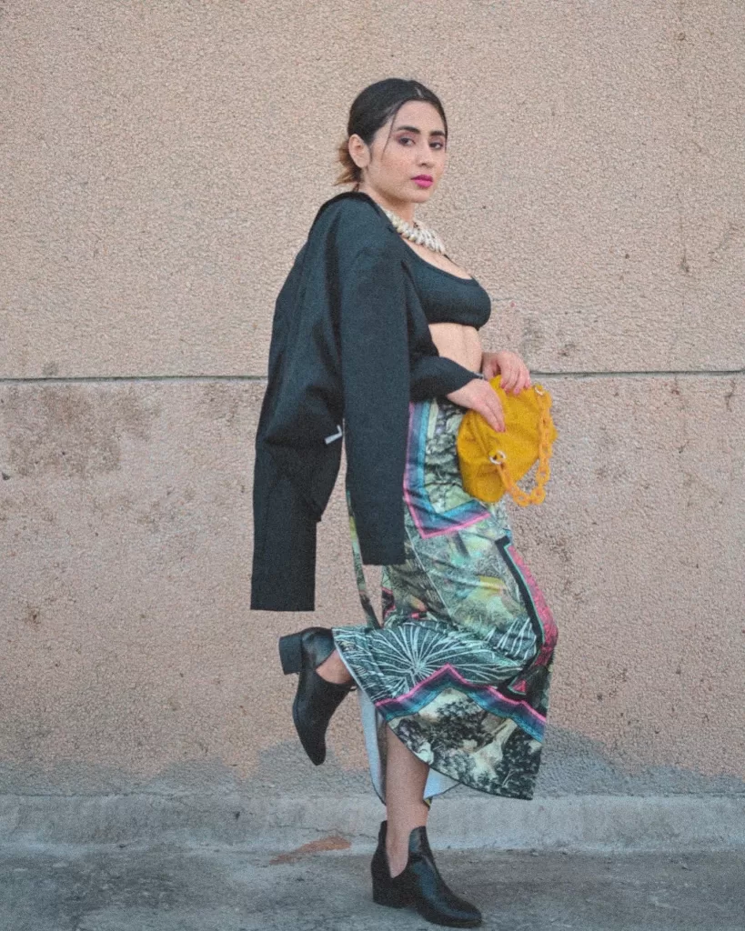 in a tailored black blazer and vibrant digital print tie-up skirt, confidently looking over her shoulder, holding a mustard-yellow bag. Paired with sleek ankle boots, her ensemble exudes a mix of power and playful chic against a neutral backdrop.
