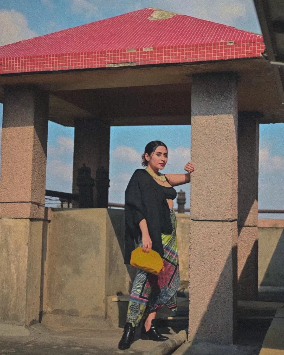 posing against a textured beige wall, donning a sharp black blazer paired with a vibrant digital print tie-up skirt. Accessorized with a bold statement necklace, holding a mustard-yellow bag, completing the look with sleek ankle boots
