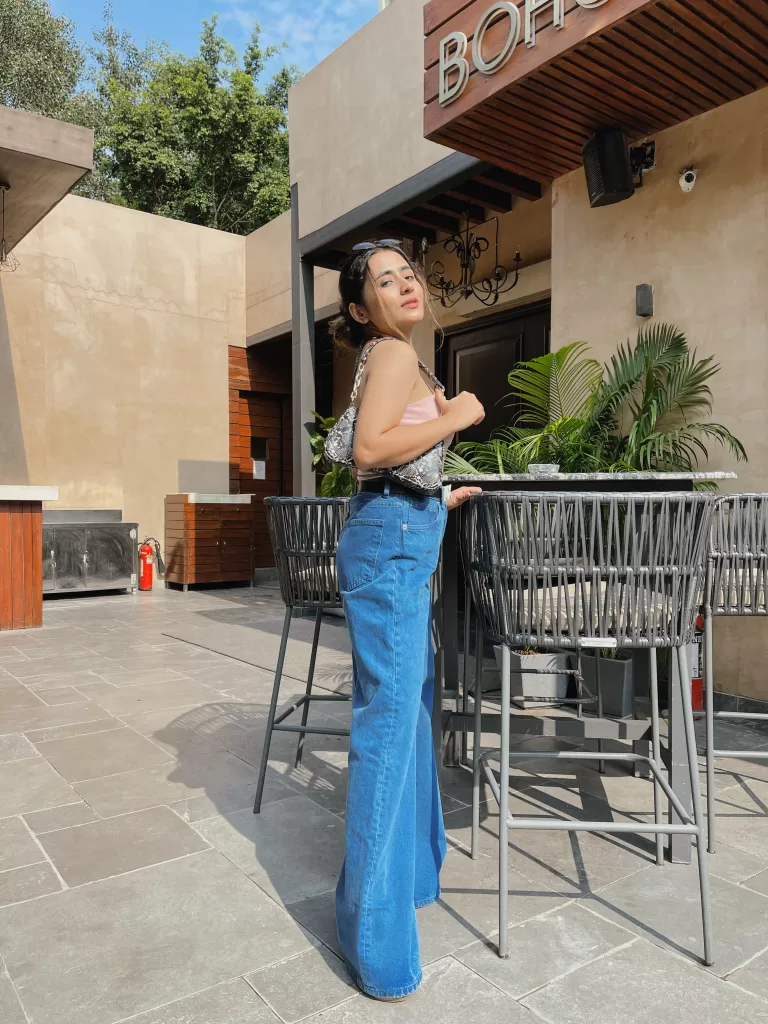 In a high-waisted jeans and a pink strapless top, accessorized with silver jewelry, striking a pose outside 'BOHCA' café amidst modern outdoor seating, with a backdrop of lush greenery and contemporary architecture.